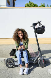 Happy young woman holding smart phone while sitting on sidewalk with electric push scooter - KIJF03138