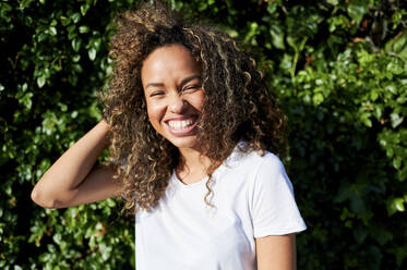 Cheerful young woman standing with hand in hair against plants during sunny day - KIJF03140