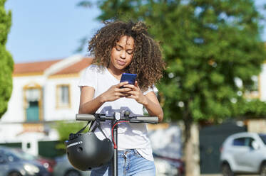 Frau, die ihr Smartphone benutzt, während sie an einem sonnigen Tag mit einem Elektroroller steht - KIJF03143