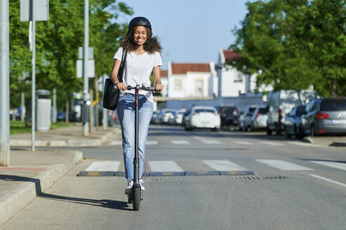 Glückliche junge Frau, die an einem sonnigen Tag auf einer Straße mit einem Elektroroller fährt - KIJF03145