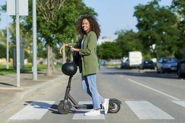 Confident young woman standing with electric push scooter on road in city - KIJF03148