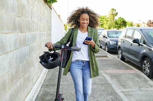 Happy young woman using smart phone while walking with electric push scooter on sidewalk - KIJF03157