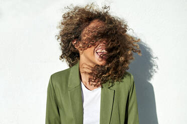 Verspielte Frau wirft ihr lockiges Haar gegen eine weiße Wand an einem sonnigen Tag - KIJF03160