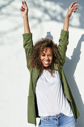 Cheerful woman standing with arms raised against white wall during sunny day - KIJF03161