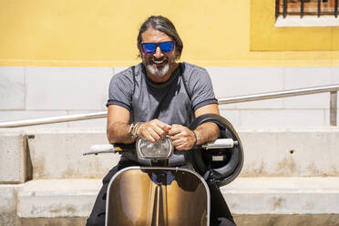 Happy mature man wearing sunglasses while sitting on motor scooter during sunny day - DLTSF00814
