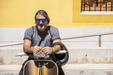 Mature man wearing sunglasses while sitting on motor scooter during sunny day - DLTSF00815