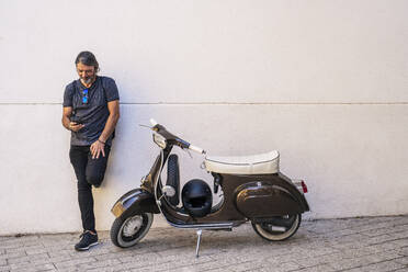 Trendy man using smart phone while standing by motor scooter against white wall - DLTSF00816