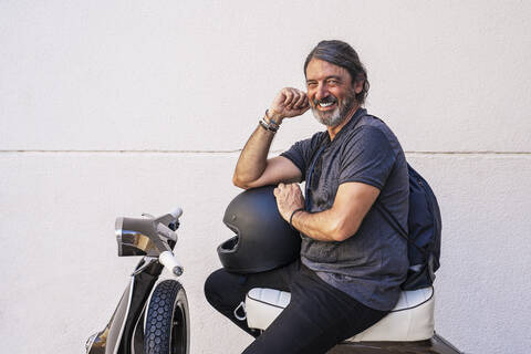 Happy man sitting on motor scooter against white wall stock photo