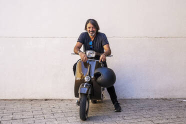 Happy man sitting on motor scooter against white wall - DLTSF00819