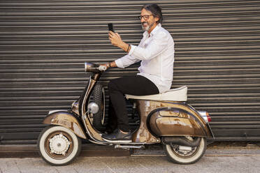 Happy mature man taking selfie while sitting on motor scooter against black shutter - DLTSF00823