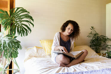 Young woman reading book while sitting on bed in log cabin - LVVF00117