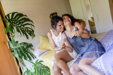 Fröhliche Frau, die ihre Freundin umarmt, während sie auf einem Bett in einem Landhaus sitzt - LVVF00133
