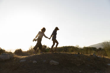 Female friends holding hands while walking on hill at countryside - LVVF00151