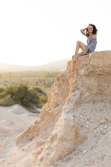 Glückliche junge Frau sitzt auf einer Klippe auf dem Lande gegen den klaren Himmel - LVVF00165
