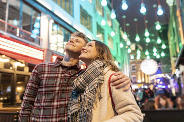 Lächelndes junges Paar, das gegen die Weihnachtsbeleuchtung in der Stadt steht und nach oben schaut - WPEF03120
