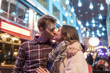 Romantisches junges Paar, das sich küsst, während es gegen die Weihnachtsbeleuchtung in der nächtlichen Stadt antritt - WPEF03122