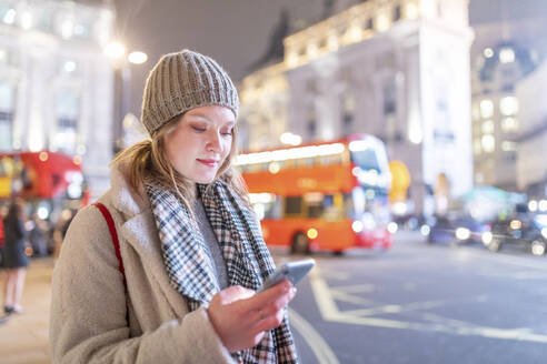 Young woman using smart phone while standing by street in Piccadilly Circus at night - WPEF03128