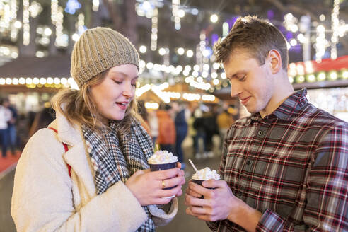 Couple holding hot chocolates while standing in Christmas market at night - WPEF03130
