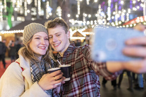 Couple holding hot chocolates taking selfie in Christmas market at night - WPEF03131