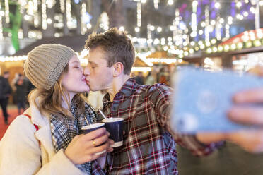 Paar hält heiße Schokolade und küsst sich, während es ein Selfie auf dem nächtlichen Weihnachtsmarkt macht - WPEF03132