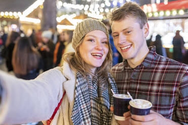 Happy couple holding hot chocolates while standing in illuminated Christmas market at night - WPEF03133