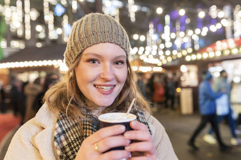 Close-up of happy beautiful woman holding hot chocolate in Christmas market at night - WPEF03134