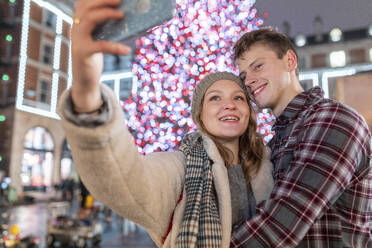 Couple taking selfie while standing against illuminated Christmas tree in city - WPEF03140