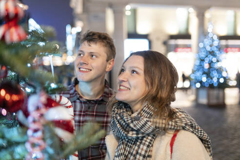 Happy young couple looking at Christmas tree and lights while standing in city - WPEF03141