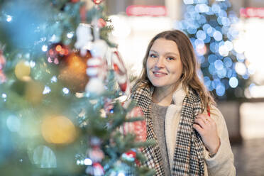 Lächelnde schöne Frau, die an einem beleuchteten Weihnachtsbaum in einer nächtlichen Stadt steht - WPEF03146