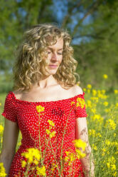 Mature woman wearing red dress with eyes closed standing amidst oilseed rapes - BFRF02253