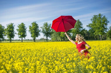Happy woman holding red umbrella while standing amidst oilseed rapes - BFRF02256