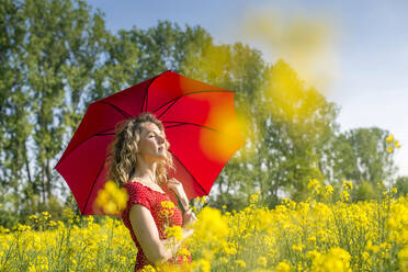 Woman wearing red dress holding umbrella while standing amidst oilseed rapes - BFRF02261