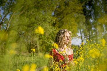 Thoughtful beautiful woman with curly hair standing amidst oilseed rapes - BFRF02262