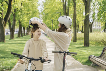 Mother wearing helmet to her daughter while standing against trees in park - AHSF02781