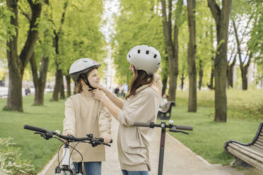 Mother wearing helmet to daughter while standing against trees in city park - AHSF02782