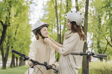 Mother wearing helmet to her daughter against trees in city park - AHSF02783