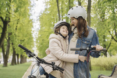 Mädchen mit Fahrrad, das seine Mutter umarmt, während es an einem Baum im Stadtpark steht - AHSF02784