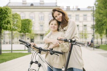 Smiling mother embracing daughter standing by scooter and bicycle in city park - AHSF02787