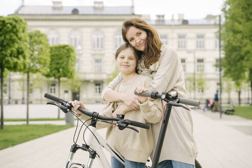 Lächelnde Mutter, die ihre Tochter umarmt, die neben einem Motorroller und einem Fahrrad im Stadtpark steht - AHSF02787