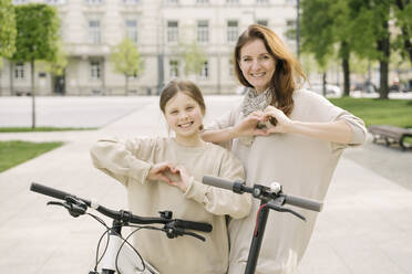 Mother with daughter gesturing heart shapes while standing by bicycle and scooter in city park - AHSF02788