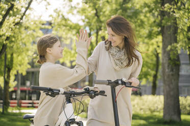 Happy mother and daughter giving high five while standing against trees in city park - AHSF02792