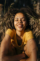 Nahaufnahme einer lächelnden jungen Frau mit Afro-Haar, die im Wald sitzt - TCEF00869