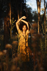 Young woman with arms raised standing amidst plants in forest during sunset - TCEF00876