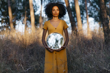 Young woman with afro hair holding mirror while standing in forest at sunset - TCEF00879