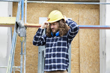 Construction worker with long hair wearing helmet while standing at construction site - MJFKF00389