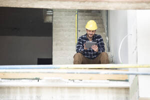 Bauarbeiter, der ein digitales Tablet benutzt, während er auf einer Baustelle auf den Stufen sitzt - MJFKF00392