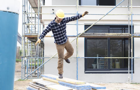 Bauarbeiter mit ausgestreckten Armen, der auf Holz gegen ein Haus läuft, lizenzfreies Stockfoto
