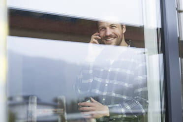 Bauarbeiter, der über sein Smartphone spricht, während er auf der Baustelle am Fenster steht - MJFKF00400