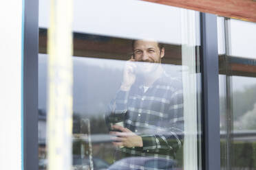 Construction worker talking over smart phone in house seen through window - MJFKF00401