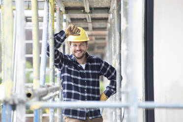 Lächelnder Bauarbeiter mit Helm an einem Baugerüst auf einer Baustelle - MJFKF00402